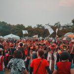 A wide-angle view of a music festival main stage at golden hour with a small group of professional photographers in dark clothing visible in the photo pit at the front, silhouetted against the bright stage and a vast crowd behind.
