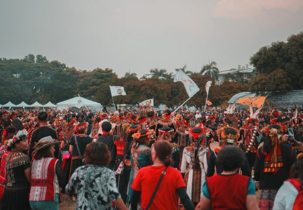 A wide-angle view of a music festival main stage at golden hour with a small group of professional photographers in dark clothing visible in the photo pit at the front, silhouetted against the bright stage and a vast crowd behind.