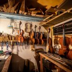 A vintage upright piano partially disassembled in a restoration workshop with the action mechanism removed and laid on a padded bench beside it, hammers and dampers visible alongside spools of new piano wire.