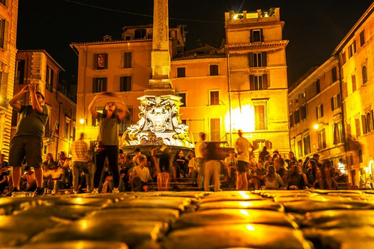 A small group of Italian folk musicians playing tambourines, accordion and violin in a stone village square at dusk with warm festoon lights strung overhead and a small crowd visible in soft focus around them.