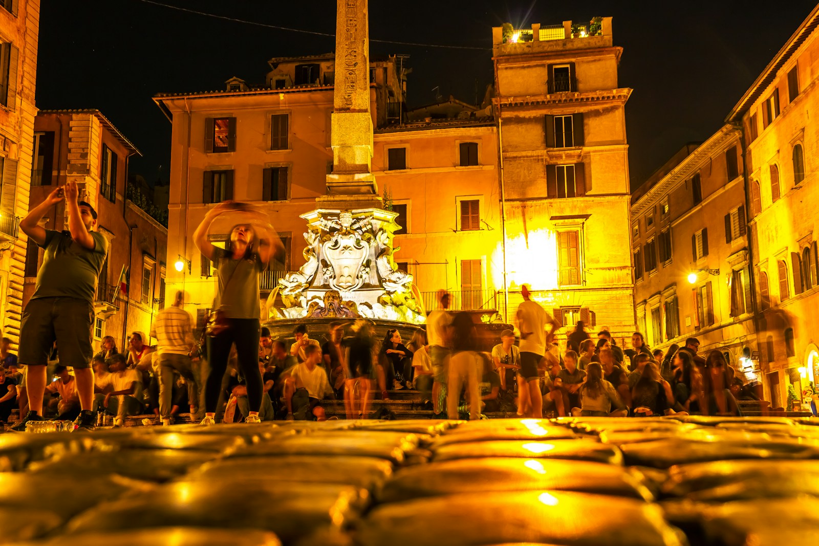 A small group of Italian folk musicians playing tambourines, accordion and violin in a stone village square at dusk with warm festoon lights strung overhead and a small crowd visible in soft focus around them.