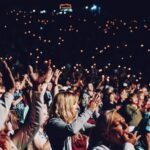 Pubblico di spalle davanti a un palco illuminato durante un concerto in una sala grande