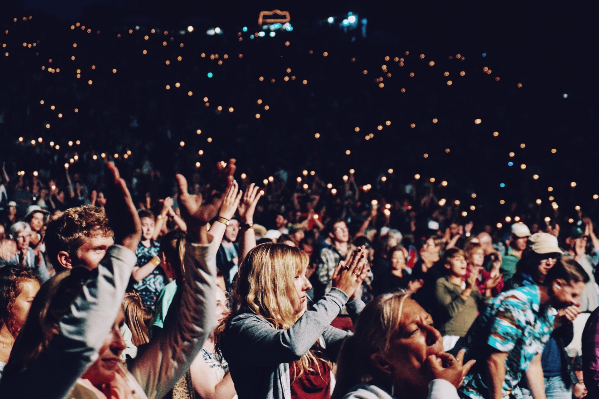 Pubblico di spalle davanti a un palco illuminato durante un concerto in una sala grande